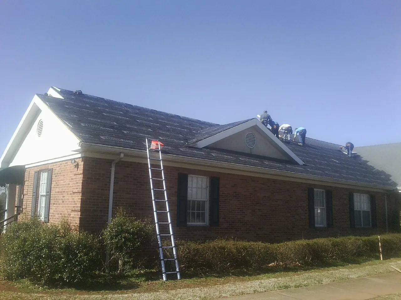 Crew tearing shingles off a brick ranch roof with a ladder set — Emergency Roof Repair work by our crew in Northwood